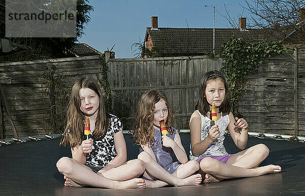 drei Mädchen essen Eis am Stiel auf einem Trampolin in Woking - England