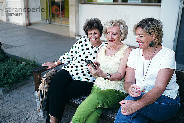 Reife Frauen lächelnd am Handy sitzend auf einer Bank