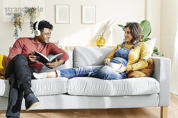 Schwangere Frau beim Essen auf der Couch mit ihrem Mann  der Musik hört und ein Buch liest. Interracial Paar Konzept