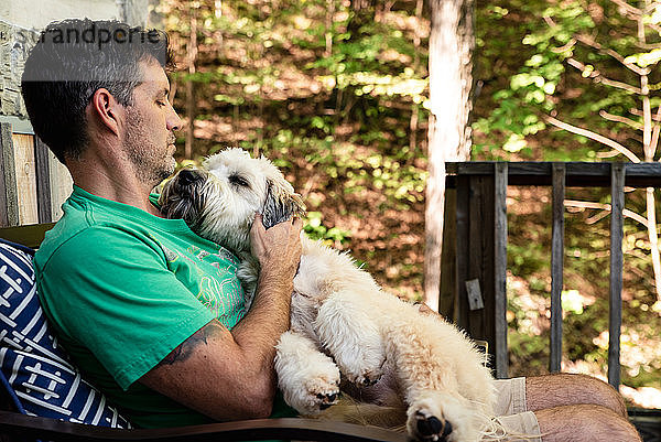 Mann  der einen flauschigen Hund auf seinem Schoß hält  draußen auf einer Terrasse im Wald.