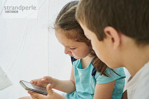 Junge und Mädchen schauen auf ein Smartphone in einem weißen Tipi-Zelt in ihrem Haus. Technologie-Konzept