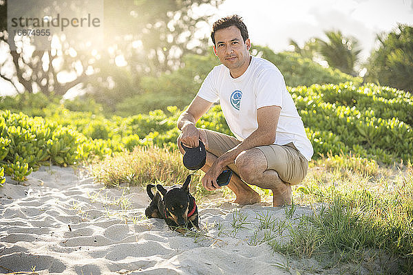 Junger fitter Mann hockt am Strand mit seinem kleinen schwarzen Hund in Hawaii