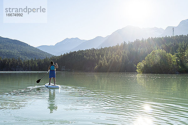 Barfüßige Frau in Kleid und Schwimmweste auf einem Paddelbrett auf einem ruhigen See an einem sonnigen Sommertag in den Bergen in British Columbia  Kanada