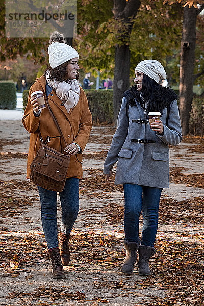 Beste Freunde plaudern im Herbst im Park mit ihrem Kaffee zum Mitnehmen. Sie tragen einen lässigen Mantel und eine Wollmütze.