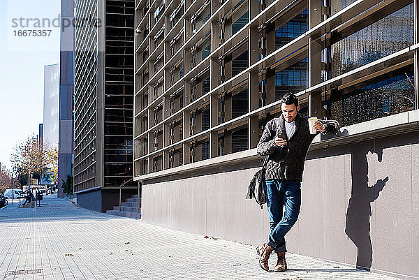 Mann mit Smartphone und Kaffee in der Hand  neben einem Bürogebäude stehend