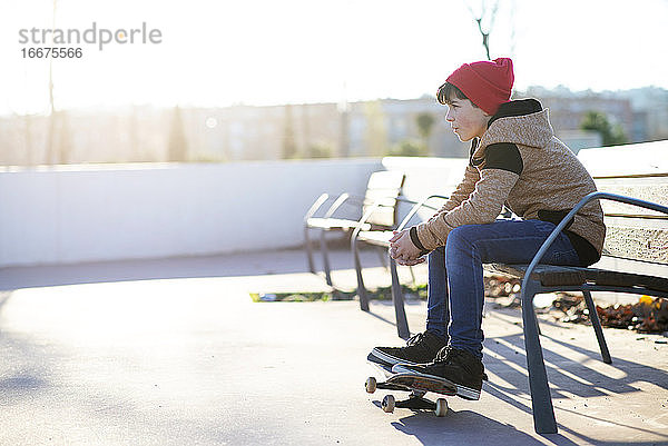 Junger Junge mit Kapuze sitzt auf einer Bank mit Schuhen über dem Skateboard