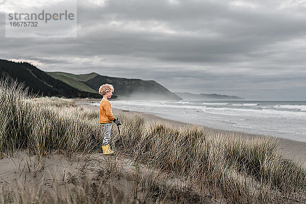 Blonder Junge mit lockigen Haaren beobachtet das Meer an einem bewölkten Tag in Neuseeland