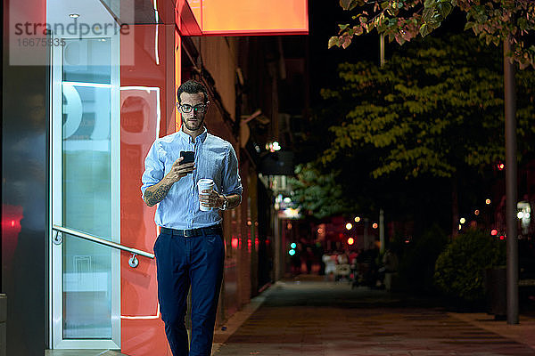 Junger Geschäftsmann spaziert mit seinem Smartphone und einer Kaffeetasse durch die Stadt.