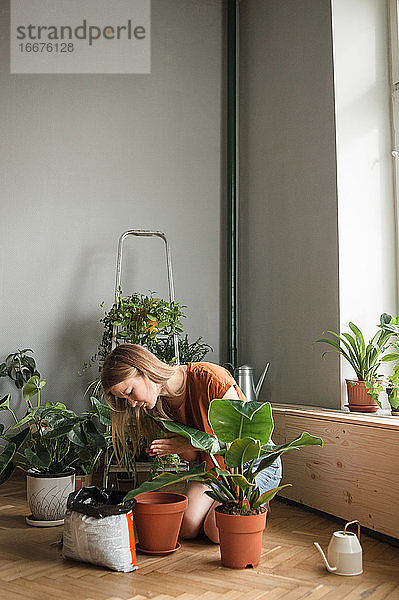 Frau schüttet Erde in einen Topf  während sie zu Hause kniend von Pflanzen umgeben ist