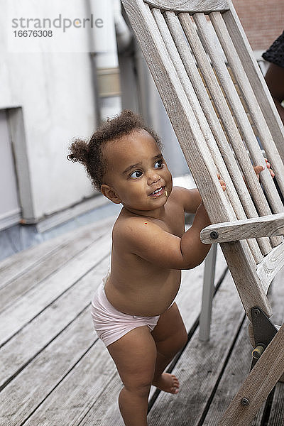 Ein nacktes Baby  das schelmisch um die Ecke eines Terrassenstuhls blickt