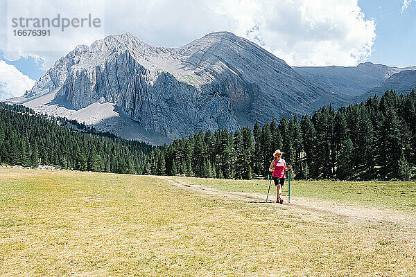 Wanderer Frau trägt Strohhut  Shorts und Rucksack auf dem Weg über eine Ebene zu Fuß mit erstaunlichen Berge im Hintergrund  während genießt die natürliche Umgebung um. Horizontal Foto.