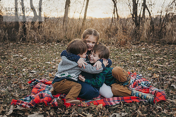 Drei Geschwister sitzen auf einer rotkarierten Decke und umarmen sich bei Sonnenuntergang