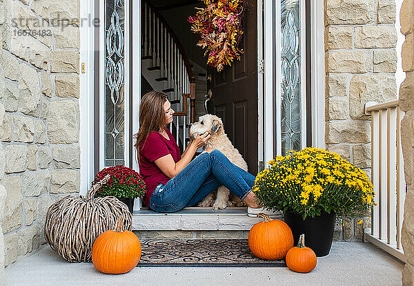 Frau und ihr Hund sitzen in der Tür eines herbstlich dekorierten Hauses.