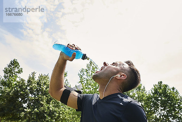 Mann trinkt nach dem Training ein Sportgetränk. Er befindet sich in einem Park im Freien.