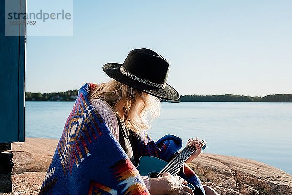 Frau sitzt auf einem Felsen und spielt Gitarre an einem sonnigen Tag am Wasser