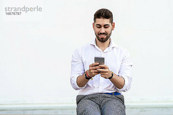 Junger bärtiger Mann sitzt an einer weißen Wand und benutzt ein Mobiltelefon