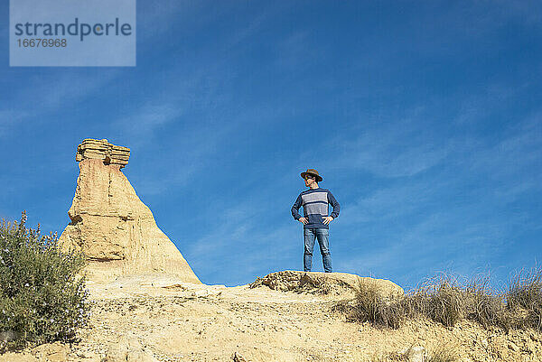 Vorderansicht eines Mannes mit Hut und Sonnenbrille  der auf Wüstendünen vor einem Felsgipfel steht