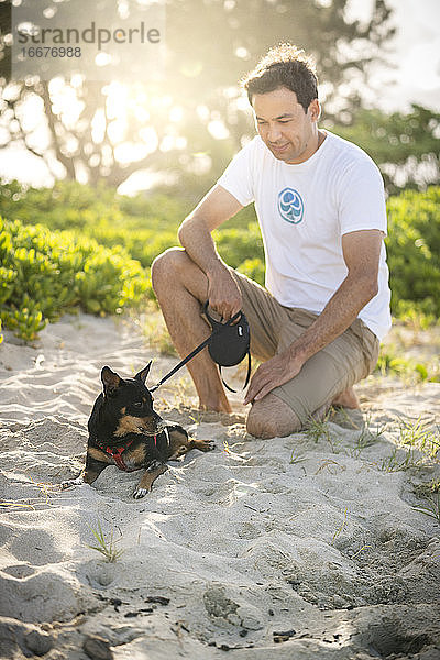 Junger fitter Mann hockt am Strand mit seinem kleinen schwarzen Hund in Hawaii