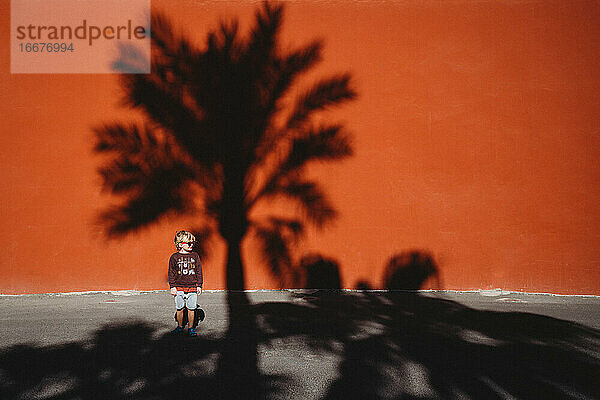 Junge steht vor einer roten Wand mit dem Schatten einer Palme