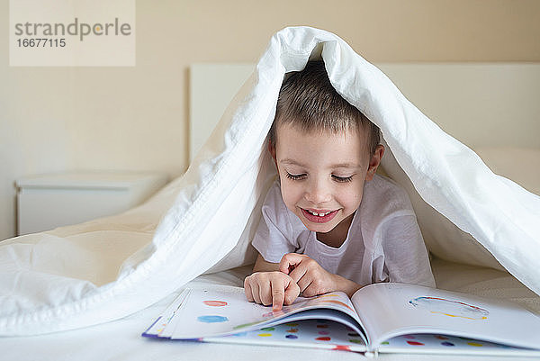Entzückendes lächelndes Kind  das unter einer Decke auf dem Bett liegt und ein Buch liest