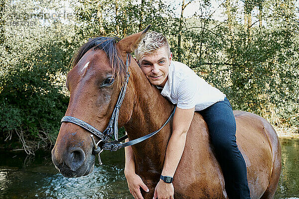 Porträt eines jungen blonden Mannes  der auf einem Pferd über einen Fluss reitet