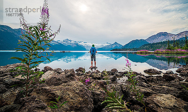 Männlicher Wanderer steht auf einem Felsen mit Blick auf einen See und verschneite Berge