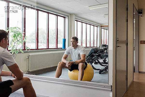 Mann sitzt im Fitnessstudio auf einem Fitnessball und betrachtet sich im Spiegel