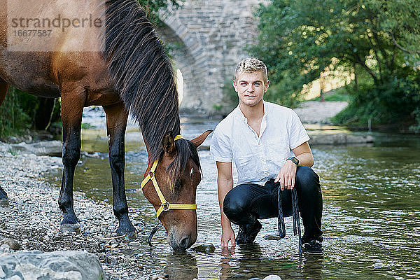 Porträt eines blonden jungen Mannes mit einem Pferd an einem Fluss