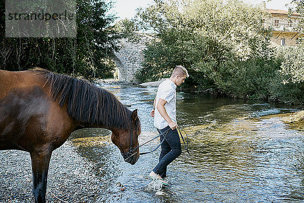 Porträt eines jungen blonden Mannes  der auf einem Pferd über einen Fluss reitet