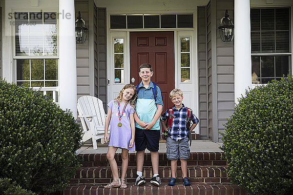 Drei lächelnde glückliche Geschwister mit Rucksäcken stehen auf Ziegelstein Front Steps