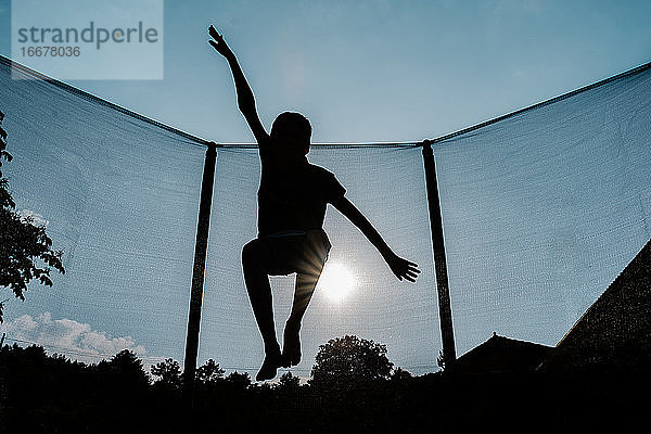 horizontales Gegenlichtfoto einer Silhouette eines barfuß springenden oder fliegenden Jungen auf einem Trampolin mit Netz und der Sonne im Hintergrund  die Sonnenstrahlen auf seinem Schatten reflektiert