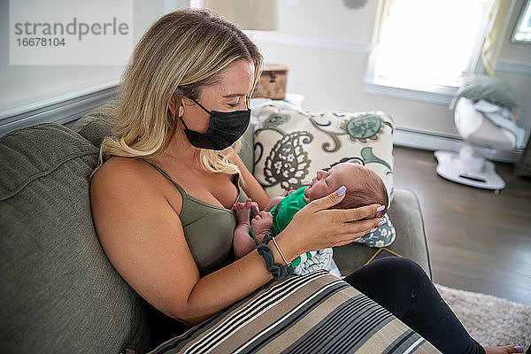 Blonde Frau mit Gesichtsmaske hält ein schläfriges Neugeborenes.