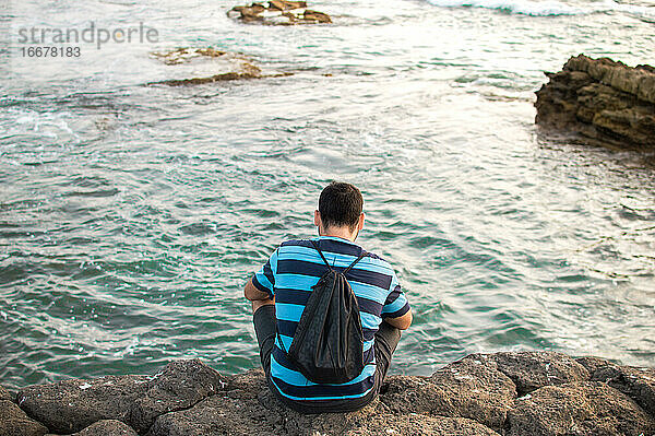 Junger Mann sitzt auf einer Klippe vor dem Meer bei Sonnenuntergang.