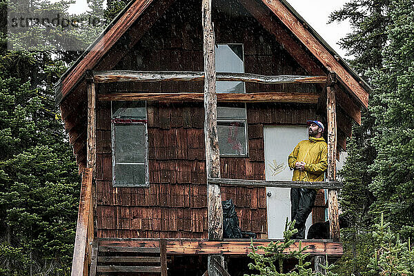 Niedriger Winkel eines männlichen Wanderers  der sich an eine verlassene Holzhütte im Wald lehnt  während er sich beim Trekking in British Columbia entspannt und wegschaut