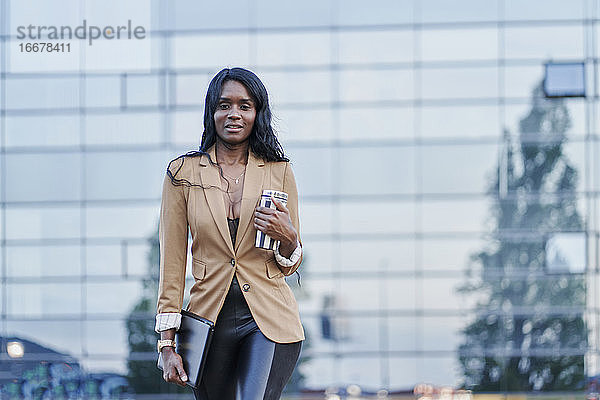 Vorderansicht Porträt einer schwarzen Frau in einem braunen Anzug  die telefoniert und einen Behälter mit Kaffee in der Hand hält