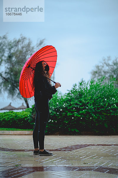 Frau mit Maske  die einen roten Regenschirm hält  schaut bei Regen auf