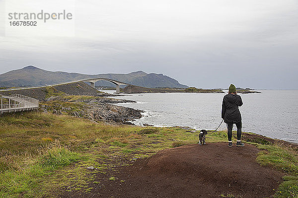 Eine Frau steht mit ihrem Hund auf einem grasbewachsenen Hügel am Meer in Norwegen