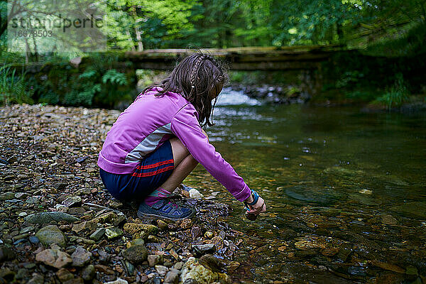 Mädchen beugt sich über ein Flussufer und spielt mit Wasser und Steinen