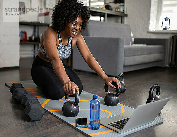 Glückliche ethnische Sportlerin mit Kettlebells und Laptop