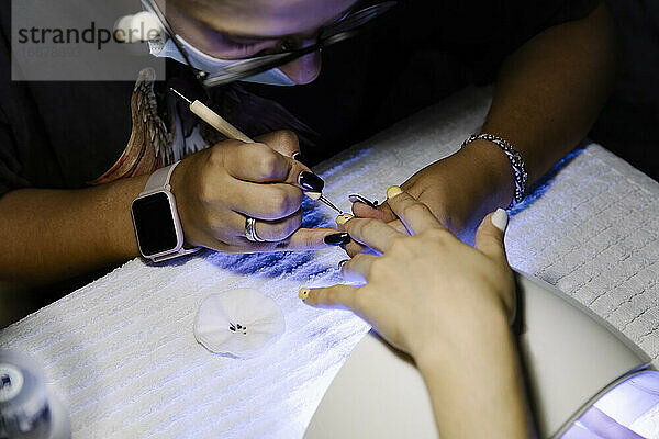 Stylistin Frau in Maske malt Punkte auf Nägel von Kunden in dunklen Salon