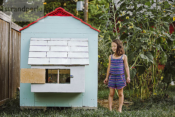 Ein glückliches kleines Mädchen steht im Hinterhof neben einem Hühnerstall und hohen Sonnenblumen