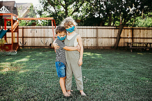 Zwei Kinder  die sich umarmen und draußen stehen und selbstgemachte Stoffmasken tragen