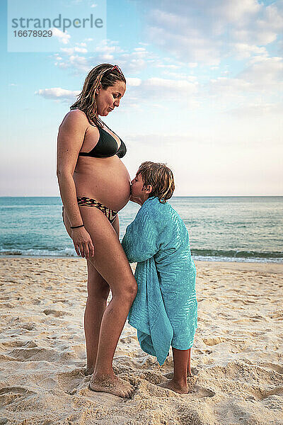 5 Jahre alter Junge küsst den Bauch seiner schwangeren Mutter am Strand