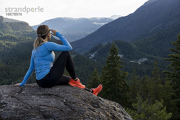 Weibliche Athletin sitzt und macht eine Pause vom Trailrunning gegen einen bewaldeten Bergrücken während eines Fitnesstrainings auf dem Lande