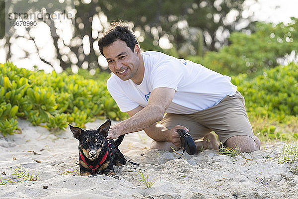 junger Mann kniet nieder  um einen schwarzen Hund zu streicheln  der am Strand mit Sand bedeckt ist