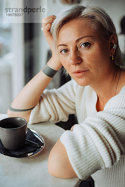 Blonde Frau sitzt am Tisch mit einer Tasse Kaffee und schaut in die Kamera