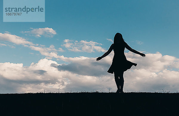 Silhouette einer Frau in einem Kleid  die gegen einen blauen Himmel mit Wolken tanzt.