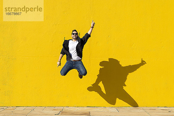 Junger Mann mit Sonnenbrille  der vor eine gelbe Wand springt.