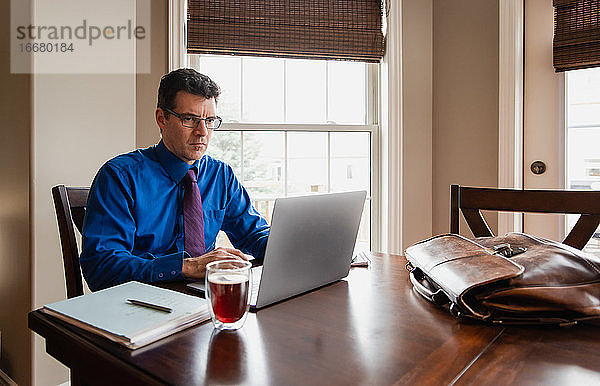 Mann in Hemd und Krawatte arbeitet von zu Hause aus am Computer am Esstisch.