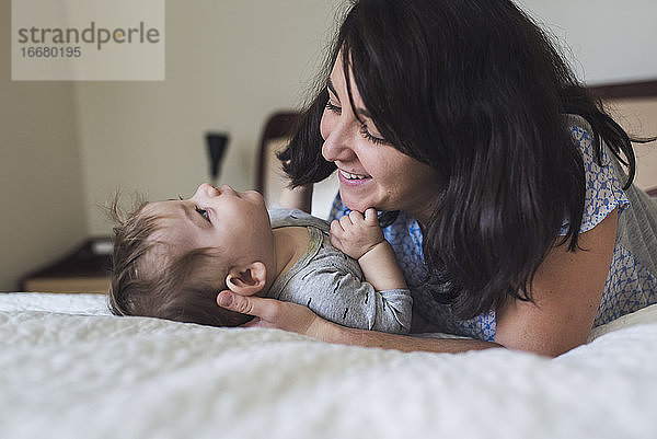 Lächelnde Mutter Mitte 30 mit dunklem Haar bewundert liebevoll das Baby auf dem Bett